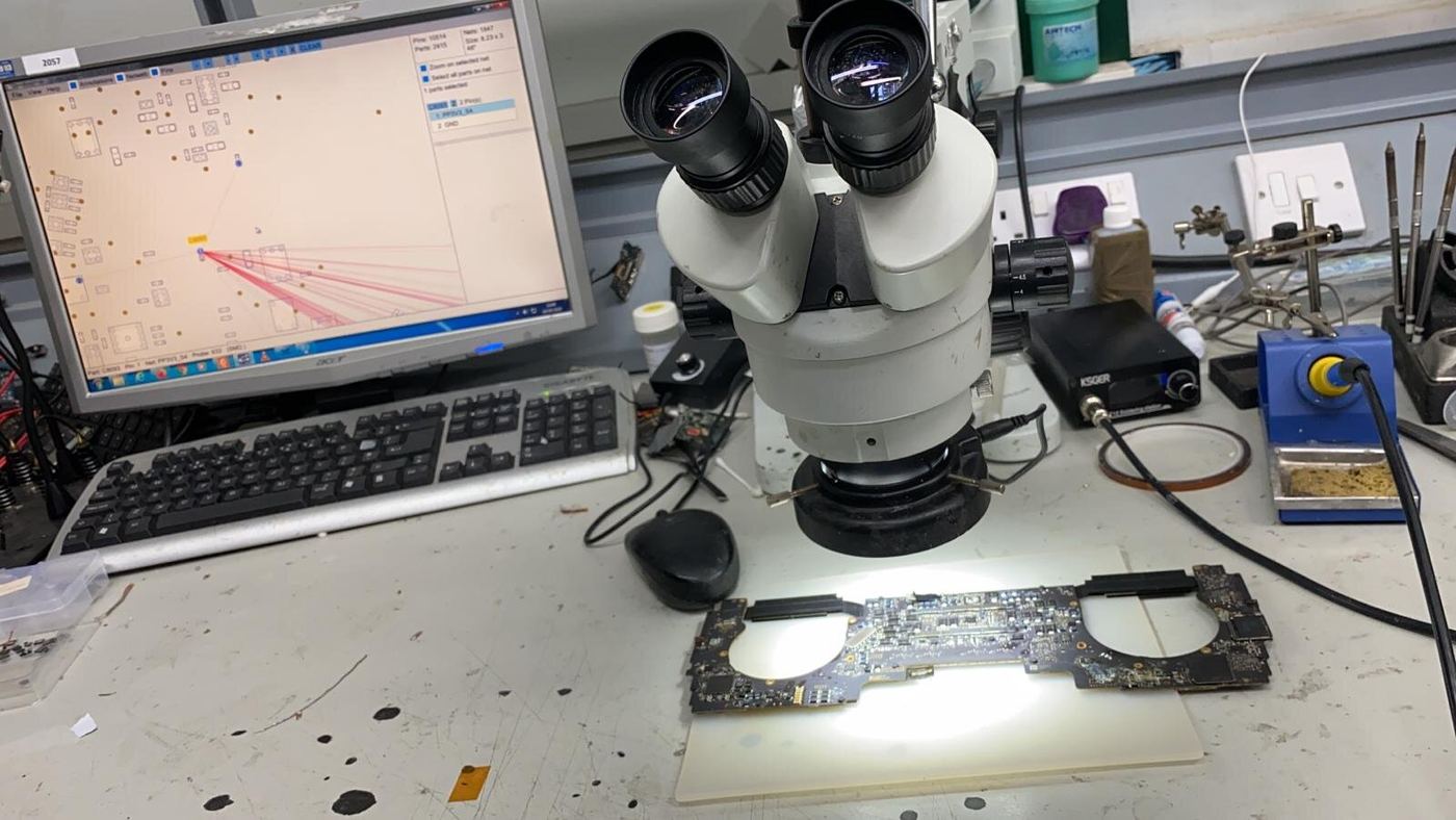 A laptop motherboard under the microscope on a repair bench, with a soldering station beside it
